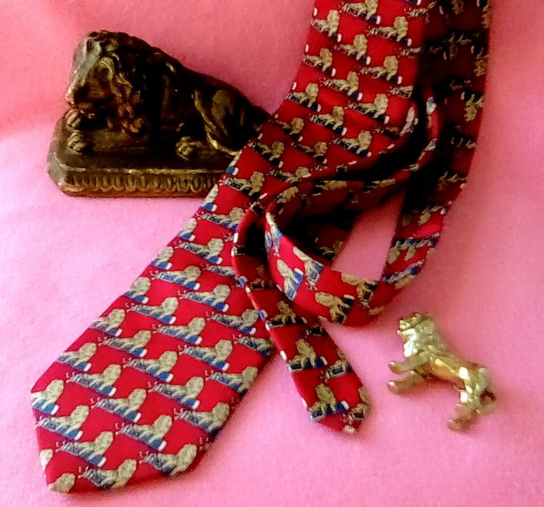 May include: A red silk tie with a repeating pattern of gold lions. The tie is lying on a pink surface with a gold lion statue.