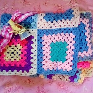 May include: A colorful crocheted blanket with a pink, blue, yellow, and green granny square pattern. A yellow bird figurine is on top of the blanket with a pink and white gingham ribbon.