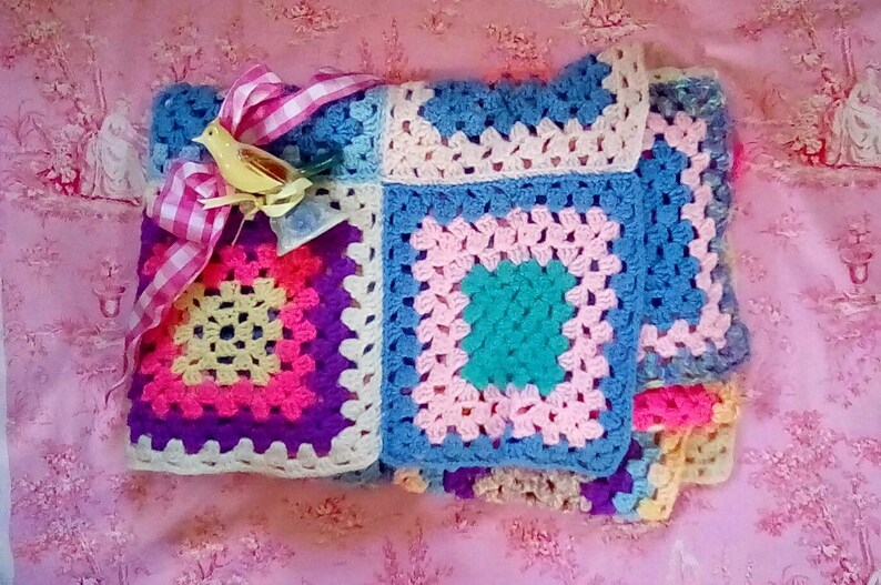 May include: A colorful crocheted blanket with a pink, blue, yellow, and green granny square pattern. A yellow bird figurine is on top of the blanket with a pink and white gingham ribbon.