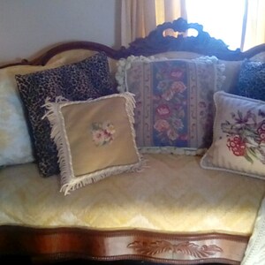 May include: A vintage-style, wooden, curved sofa with a yellow fabric upholstery. The sofa has five pillows, including a leopard print pillow, a floral pillow, and a pillow with a floral design. The sofa is decorated with a white knitted throw blanket.