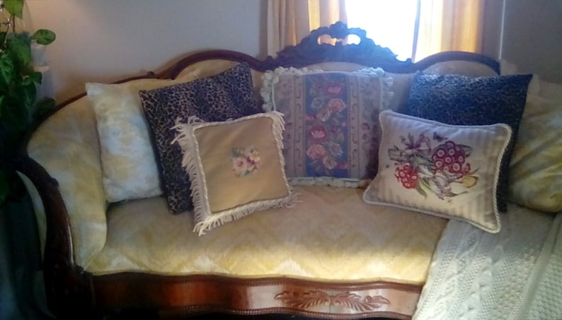 May include: A vintage-style, wooden, curved sofa with a yellow fabric upholstery. The sofa has five pillows, including a leopard print pillow, a floral pillow, and a pillow with a floral design. The sofa is decorated with a white knitted throw blanket.
