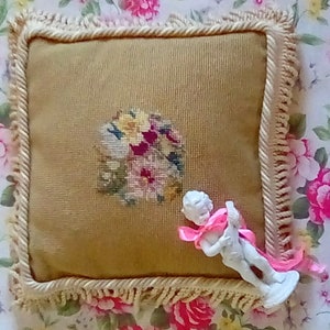 May include: A square beige pillow with a floral embroidery design and a white figurine of a cherub holding a pink ribbon on top of it. The pillow has a white fringe border.
