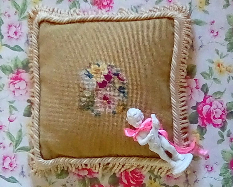 May include: A square beige pillow with a floral embroidery design and a white figurine of a cherub holding a pink ribbon on top of it. The pillow has a white fringe border.