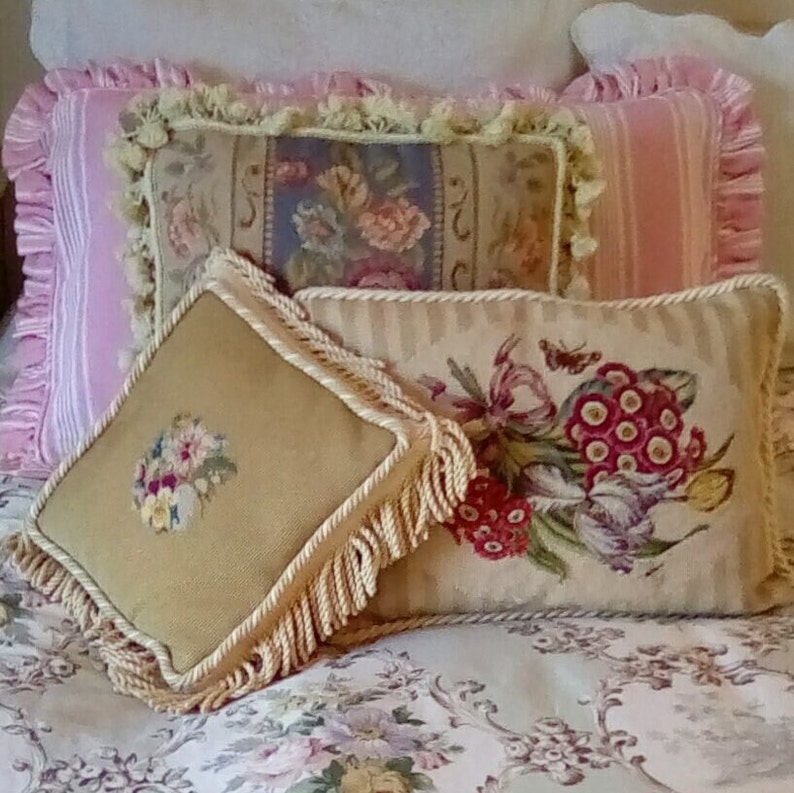 May include: Three decorative throw pillows with floral embroidery. The pillows are beige with cream colored fringe and are arranged on a floral patterned bedspread.