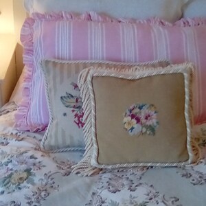 May include: A bed with a white, pink, and beige patterned bedding set. The bedding features a white pillow with a pink and white striped pillow on top, and a beige pillow with a floral embroidery on top of that. The bedspread is a light beige with a floral pattern.