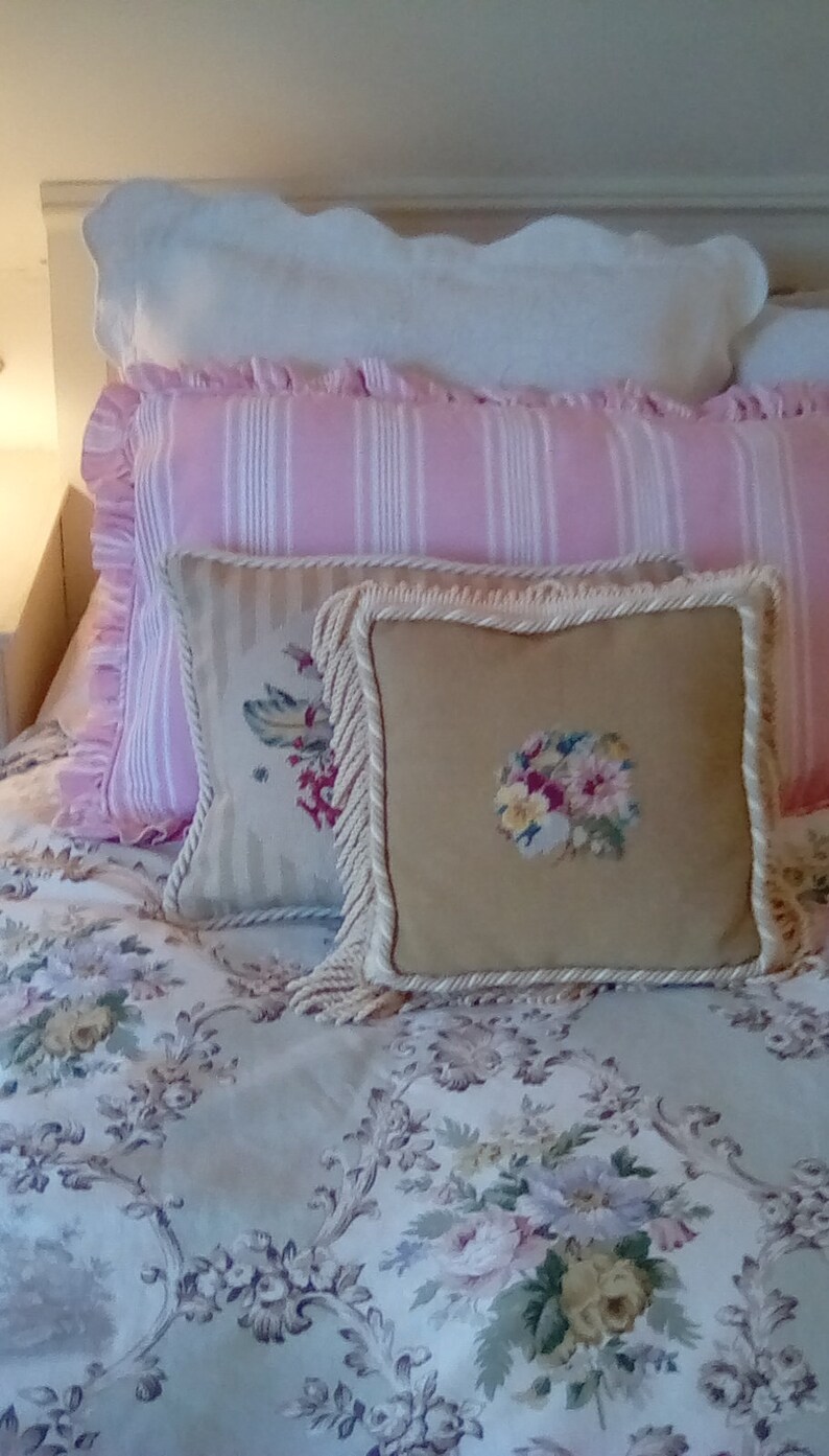 May include: A bed with a white, pink, and beige patterned bedding set. The bedding features a white pillow with a pink and white striped pillow on top, and a beige pillow with a floral embroidery on top of that. The bedspread is a light beige with a floral pattern.