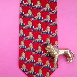 May include: A red silk tie with a repeating pattern of gold lions sitting on blue and white striped barrels. A gold lion figurine is placed on the right side of the tie.