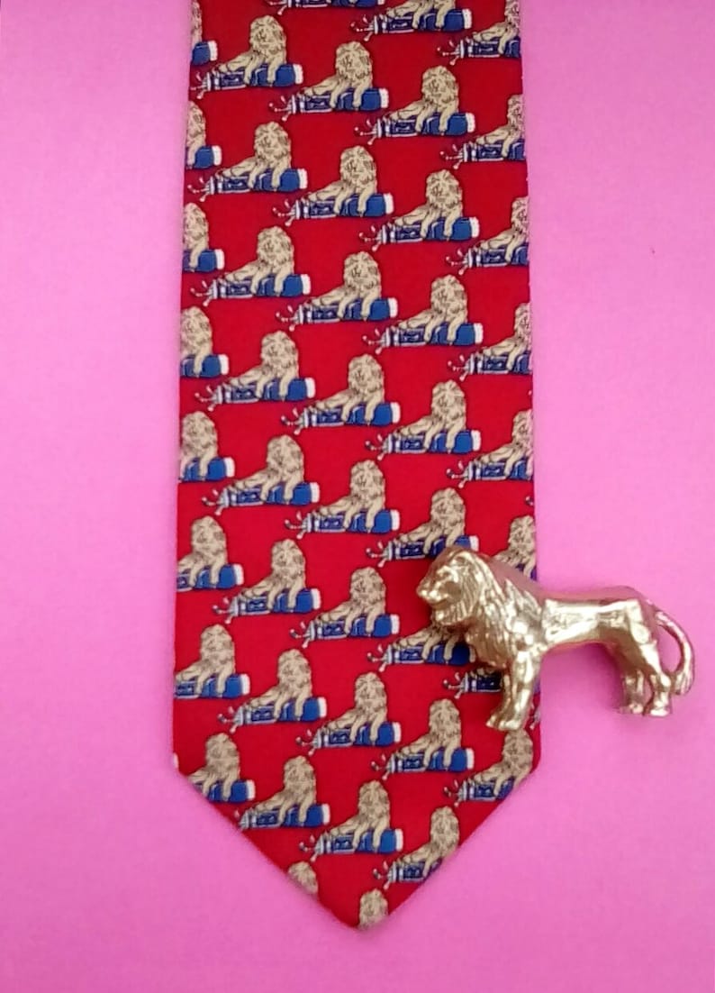 May include: A red silk tie with a repeating pattern of gold lions sitting on blue and white striped barrels. A gold lion figurine is placed on the right side of the tie.