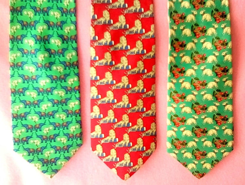 May include: Three silk neckties with different patterns. The first tie has a green background with a repeating pattern of elephants and flowers. The second tie has a red background with a repeating pattern of lions and flowers. The third tie has a green background with a repeating pattern of rabbits and flowers.