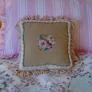 May include: A tan pillow with a floral embroidery design and a white fringe border sits on a pink and white striped pillow, which is on top of a floral patterned bedspread.