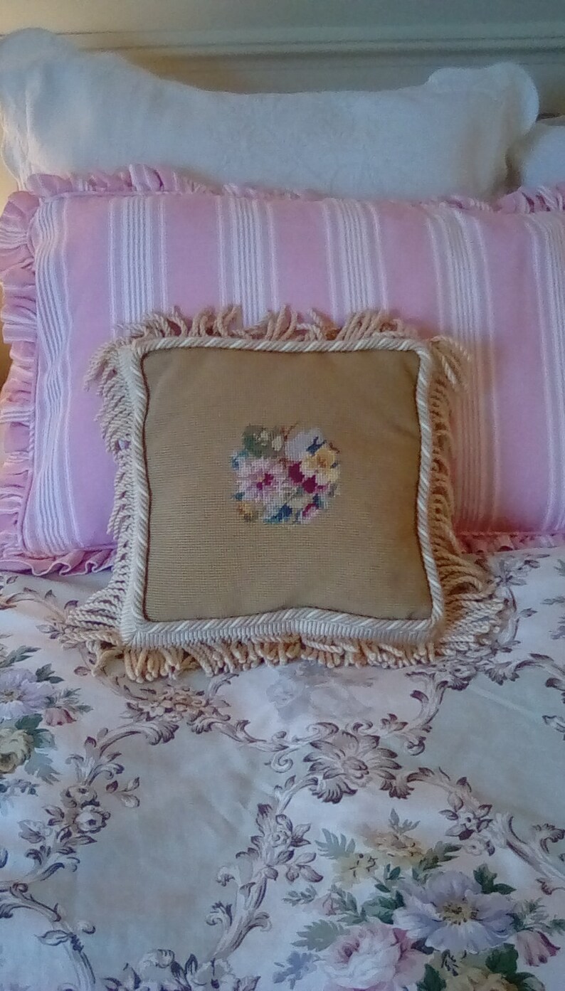 May include: A tan pillow with a floral embroidery design and a white fringe border sits on a pink and white striped pillow, which is on top of a floral patterned bedspread.