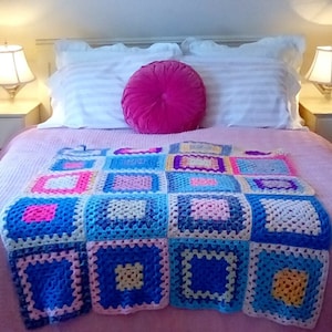 May include: A pink bed with white striped pillowcases and a pink velvet round pillow. A colorful crocheted blanket with squares of pink, blue, yellow, and white is on top of the bed.