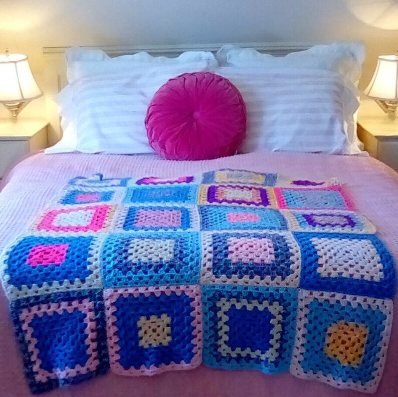 May include: A pink bed with white striped pillowcases and a pink velvet round pillow. A colorful crocheted blanket with squares of pink, blue, yellow, and white is on top of the bed.