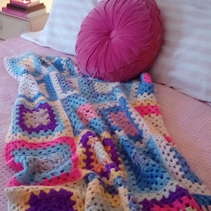 May include: A colorful crocheted blanket with squares of pink, blue, yellow, and purple. The blanket is draped over a pink bedspread with a pink velvet pillow.