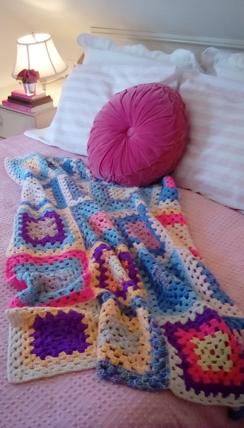 May include: A colorful crocheted blanket with squares of pink, blue, yellow, and purple. The blanket is draped over a pink bedspread with a pink velvet pillow.