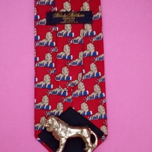 May include: Red silk tie with a repeating pattern of gold lions on blue and white barrels. The tie has a black label with the text "Brooks Brothers Makers of Fine Clothes Since 1818" and a gold lion tie tack.