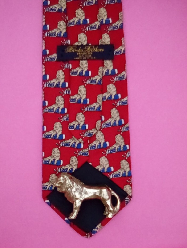 May include: Red silk tie with a repeating pattern of gold lions on blue and white barrels. The tie has a black label with the text "Brooks Brothers Makers of Fine Clothes Since 1818" and a gold lion tie tack.