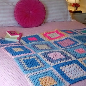May include: A pink bed with a white and pink striped pillow and a pink round pillow. A crocheted blanket with squares of blue, pink, yellow, and white is on the bed.