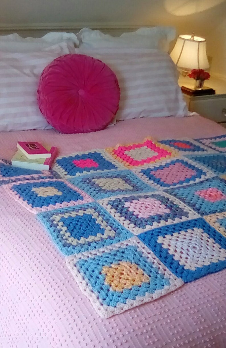 May include: A pink bed with a white and pink striped pillow and a pink round pillow. A crocheted blanket with squares of blue, pink, yellow, and white is on the bed.