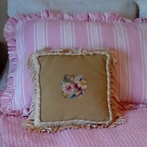 May include: A pink and white striped pillow with a cream colored pillow with a floral embroidery design on top. Both pillows have fringe trim.
