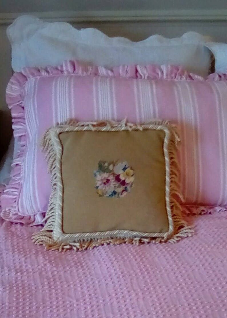 May include: A pink and white striped pillow with a cream colored pillow with a floral embroidery design on top. Both pillows have fringe trim.