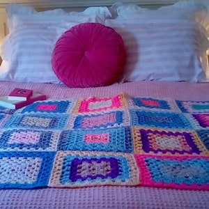 May include: A pink bed with white pillows and a pink round pillow. A crocheted blanket with squares of blue, pink, yellow, and purple is on the bed. Two books are on the blanket.