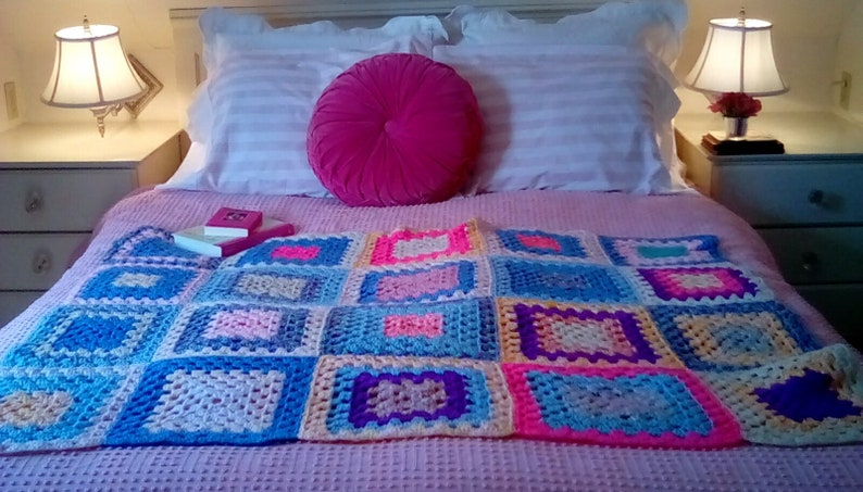 May include: A pink bed with white pillows and a pink round pillow. A crocheted blanket with squares of blue, pink, yellow, and purple is on the bed. Two books are on the blanket.