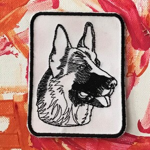 May include: Black and white embroidered patch of a German Shepherd dog's head. The dog is looking to the right with its tongue sticking out.