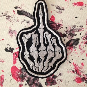 May include: A black and white embroidered patch of a skeleton middle finger. The patch is on a white background with red and black paint splatters.