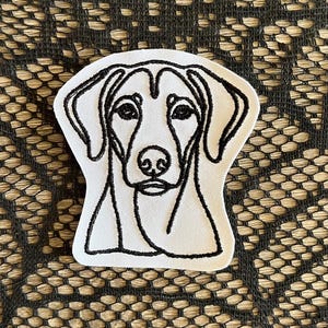 May include: A white patch featuring a black line drawing of a dog's face. The dog's head is centered, with detailed eyes, nose, and ears. The patch has a simple, clean design, suitable for various applications.