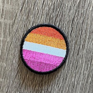 May include: A round, embroidered patch with the lesbian pride flag colors: orange, yellow, white, and pink. The patch has a black border.