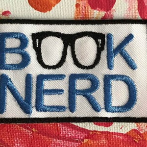 May include: White embroidered patch with black outline. The patch says "BOOK NERD" in blue embroidery with a pair of black glasses in the middle.