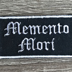 May include: Black embroidered patch with white stitching. The patch features the text "Memento Mori" in a gothic font.
