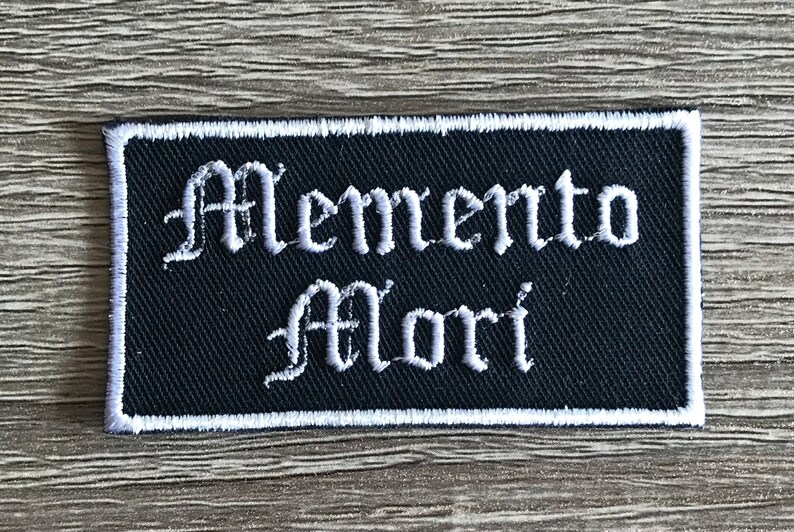 May include: Black embroidered patch with white stitching. The patch features the text "Memento Mori" in a gothic font.