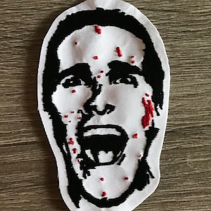 May include: A white embroidered patch with a black silhouette of a man's face screaming. The man has red blood splatters on his face.