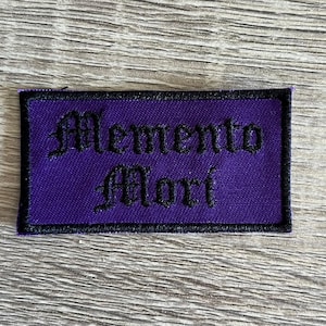May include: Purple embroidered patch with black stitching. The patch reads "Memento Mori" in a gothic font.