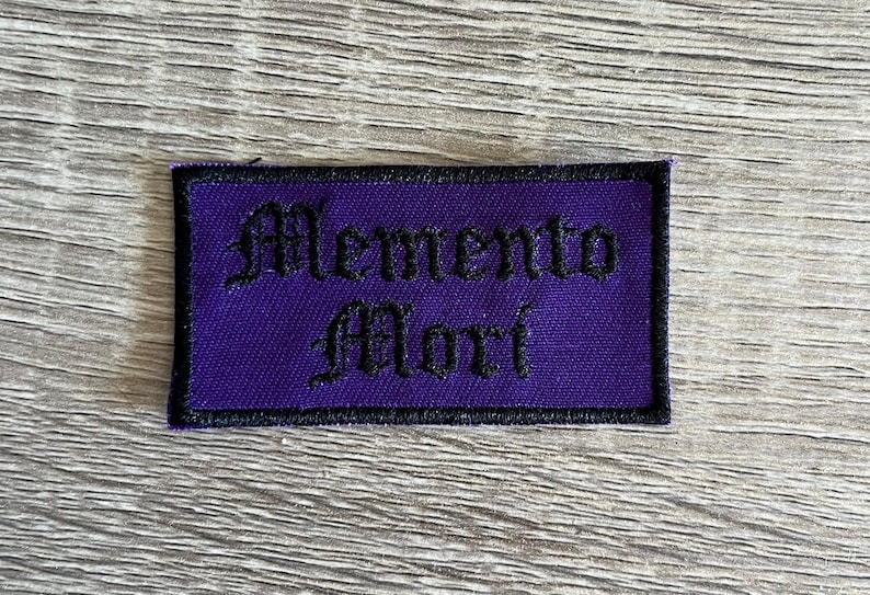 May include: Purple embroidered patch with black stitching. The patch reads "Memento Mori" in a gothic font.