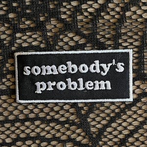 May include: A rectangular black embroidered patch with a white border and the white text somebody's problem written in a lowercase serif font. The patch is positioned against a black lace fabric background with a repeating floral pattern.