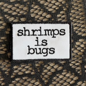 May include: A white rectangular fabric label with the words "shrimps is bugs" embroidered in black. The label is set against a textured background of interwoven dark brown and black fibers.