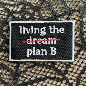 May include: A black rectangular patch with white text that reads "living the dream" with a red line through "dream" and the text "plan B" below. The patch has a white border.