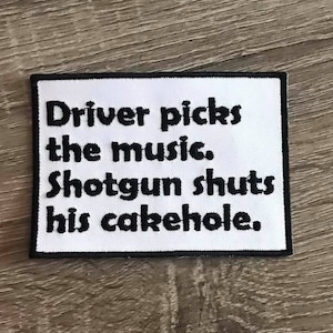 May include: A white rectangular patch with black stitched border. The patch features the text "Driver picks the music. Shotgun shuts his cakehole." in bold black letters. The patch is likely an iron-on or sew-on.