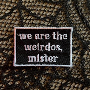 May include: A black rectangular patch with white embroidered text that reads "we are the weirdos, mister". The patch has a white border and is set against a textured background.