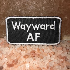 May include: A black rectangular patch with white embroidered text that reads "Wayward AF." The patch has a white border and is displayed on a bed of pink salt crystals.