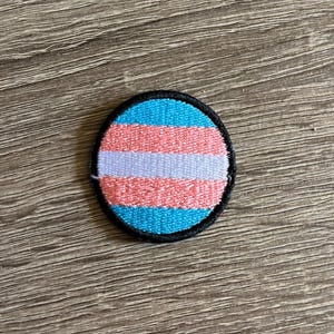 May include: A round, embroidered patch with the transgender pride flag colors: light blue, pink, and white. The patch is outlined in black.