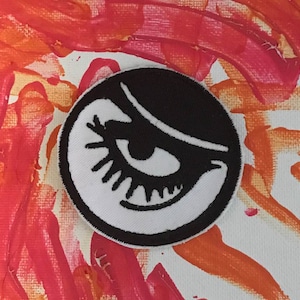 May include: A black and white embroidered patch with a large eye and eyelashes. The eye is white with a black pupil and the eyelashes are black. The patch is circular and has a black border.