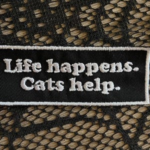 May include: A rectangular black embroidered patch with a white border. The text "Life happens. Cats help." is stitched in white thread in a serif font across the center of the patch. The design is clean, featuring high-contrast white lettering.