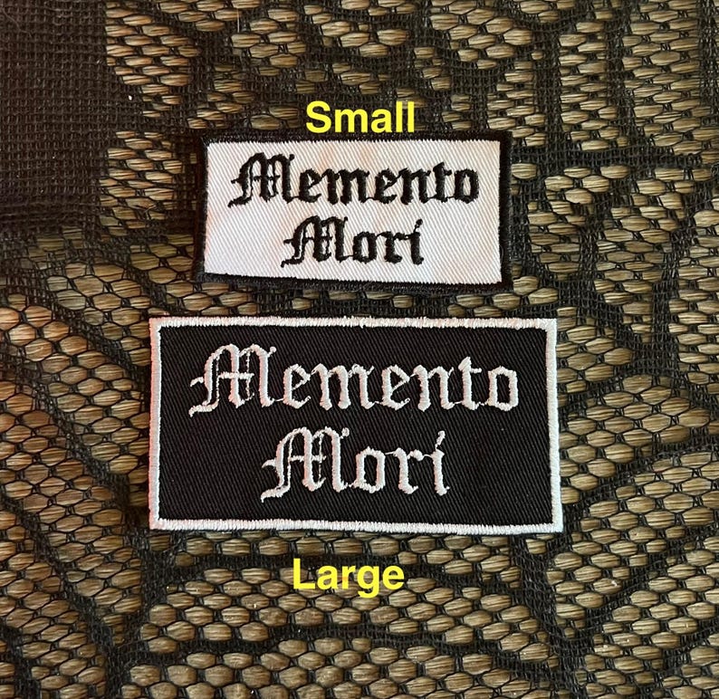 May include: Two black and white embroidered patches with the text "Memento Mori" in a gothic font. The smaller patch is on a white background and the larger patch is on a black background.