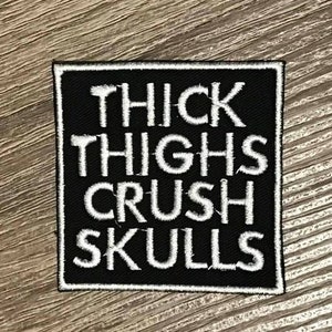 May include: Black iron-on patch with white text that reads "THICK THIGHS CRUSH SKULLS".