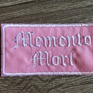 May include: A pink fabric patch with white embroidered text that reads "Memento Mori" in a gothic font.