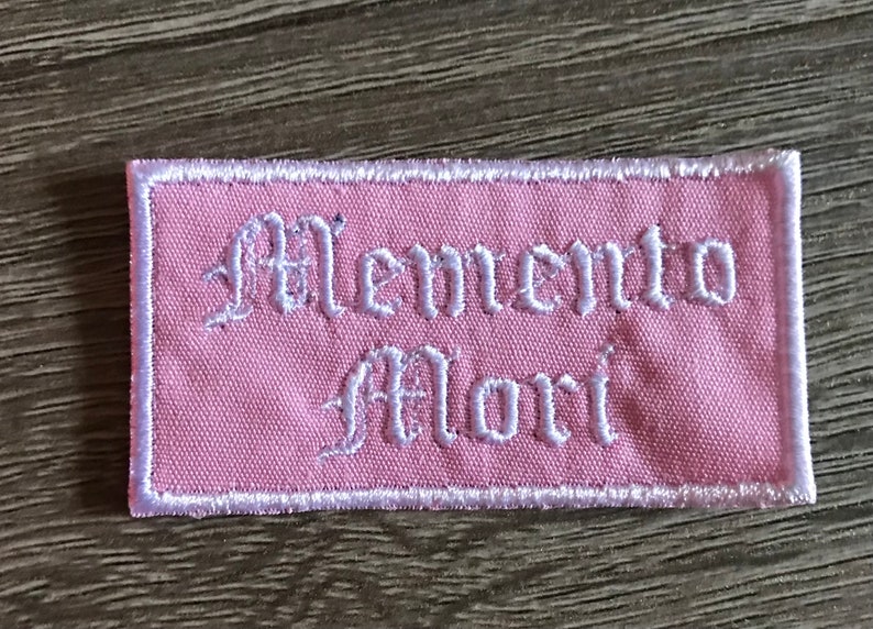 May include: A pink fabric patch with white embroidered text that reads "Memento Mori" in a gothic font.